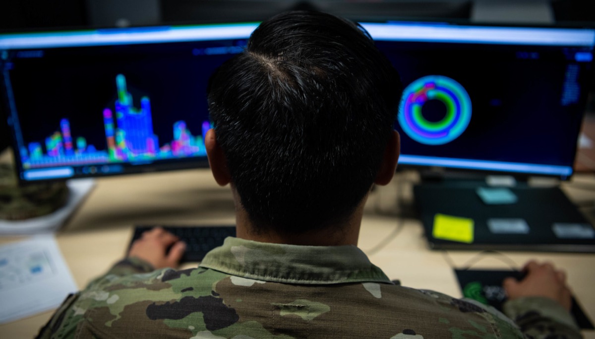 A cyber operator working across two monitors of live system telemetry.